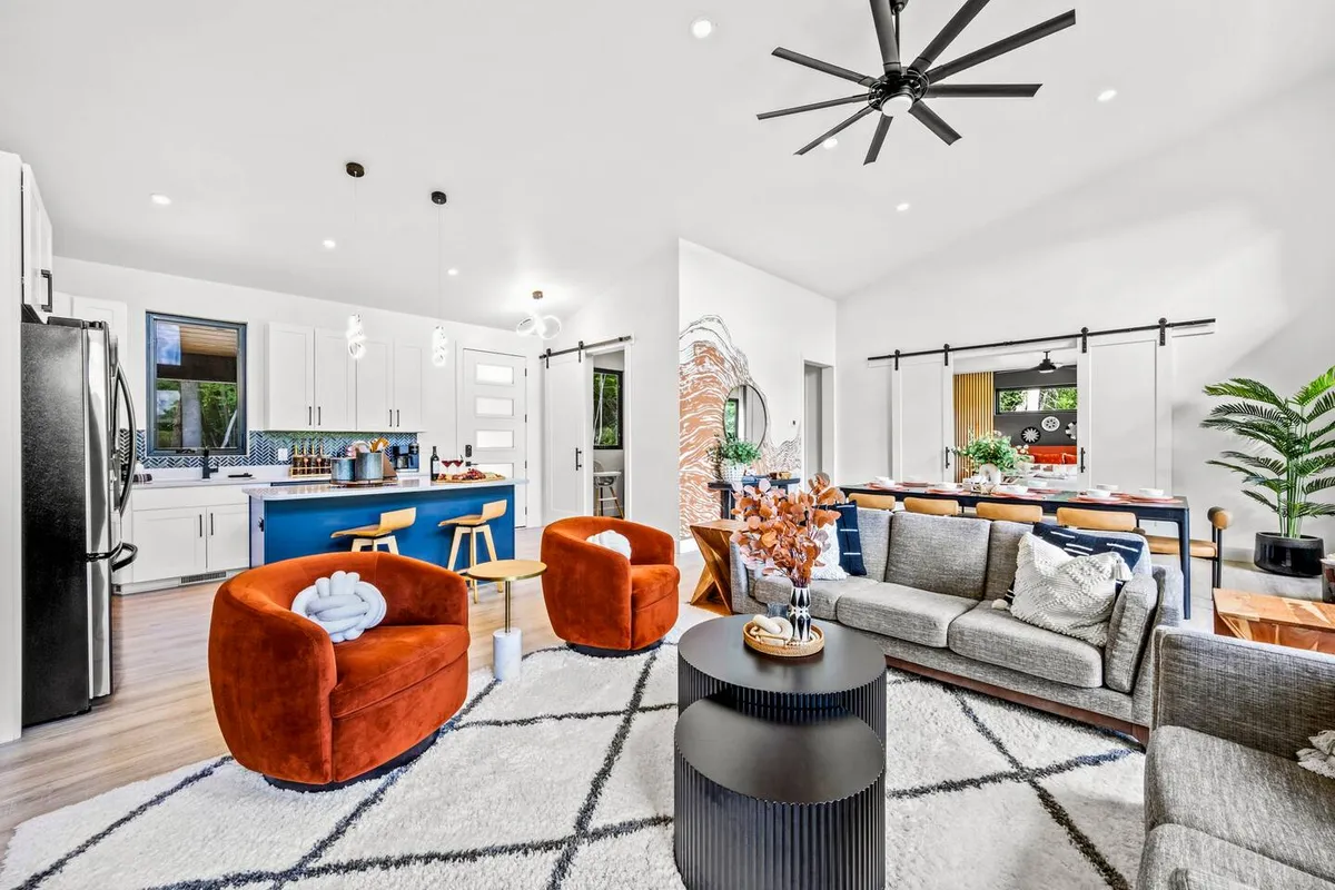 A vibrant open living and dining area with colorful orange armchairs, a grey sofa, and a blue kitchen island in the background. Decorative plants and ample natural light complete the scene, making it feel lively yet comfortable.