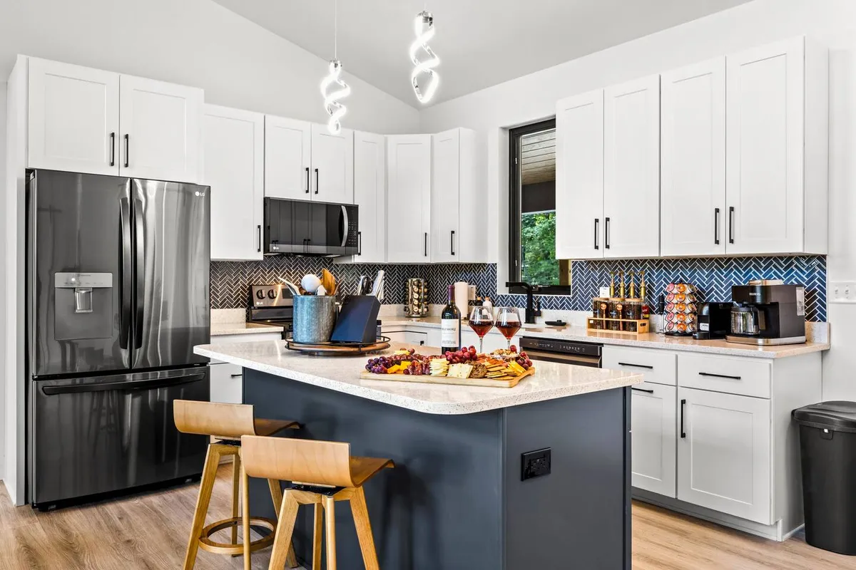 The kitchen’s island countertop is set with snacks and drinks, lit by modern fixtures and anchored by a sleek black refrigerator. The scene captures a vibrant, welcoming atmosphere where guests can gather and socialize.