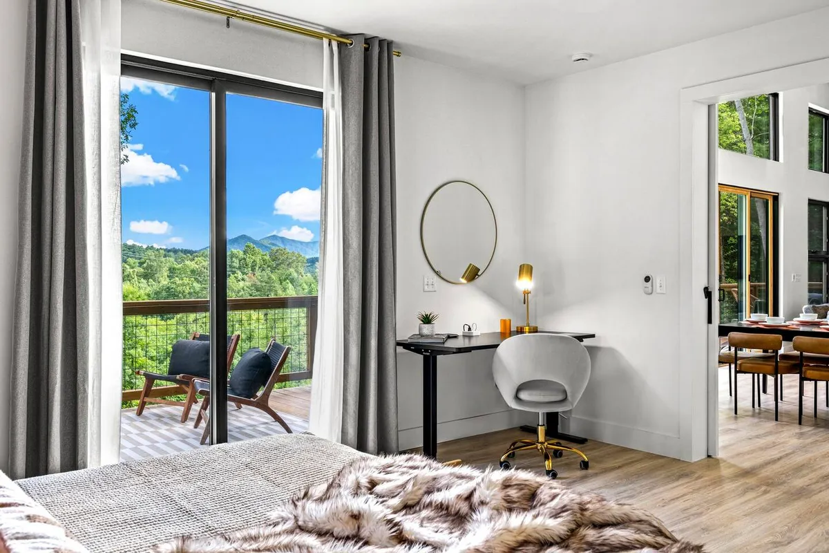 A calming bedroom with a large window offering stunning mountain views, a stylish desk and chair for remote work or journaling, and a soft bed with a luxurious fur throw. The ambiance is serene and conducive to both relaxation and productivity.