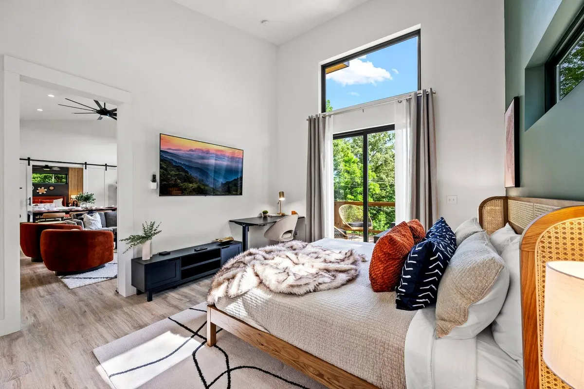 A luxurious bedroom featuring a plush bed with a soft throw, large windows framing tranquil nature views, a television displaying a scenic landscape, and a stylish desk with a chair. The atmosphere is calm and inviting, perfect for unwinding or getting some focused work done while enjoying the mountain scenery.