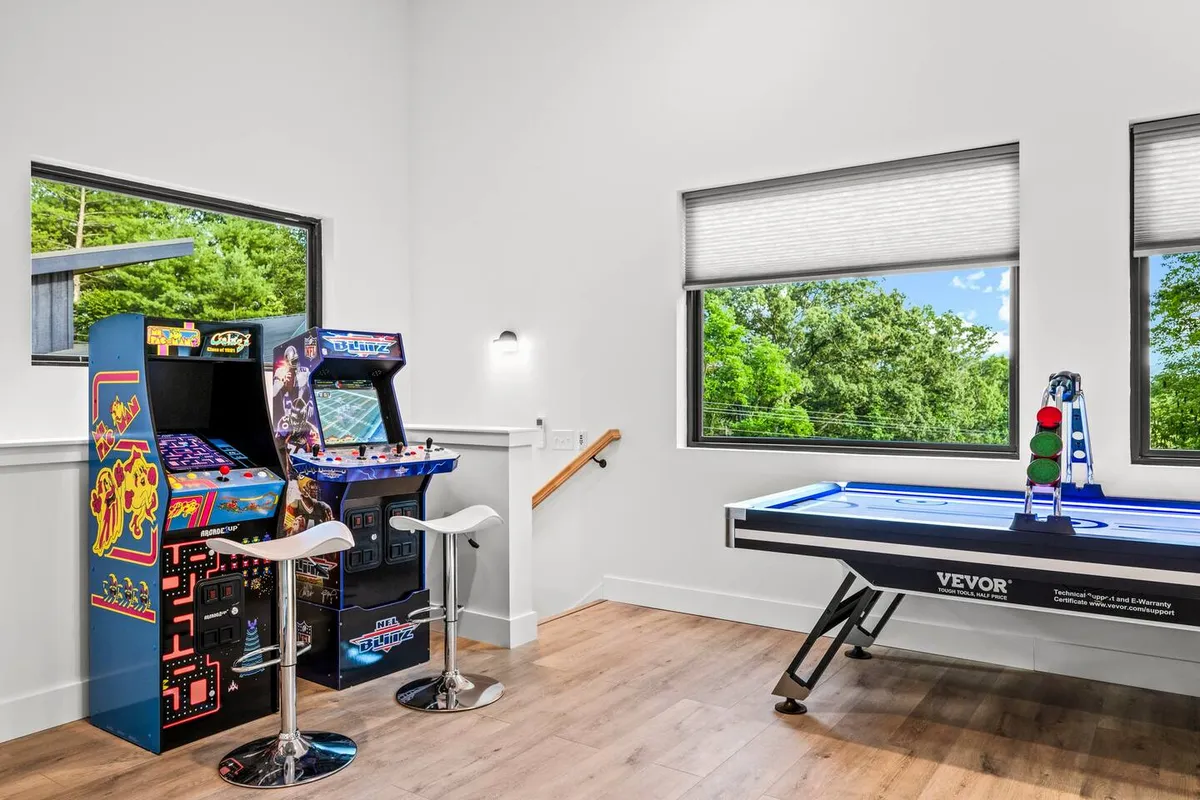 A lively game room featuring multiple arcade machines and an air hockey table, with large windows letting in natural light. Groups of friends are seen playing arcade games and enjoying air hockey, capturing a high-energy, social atmosphere.