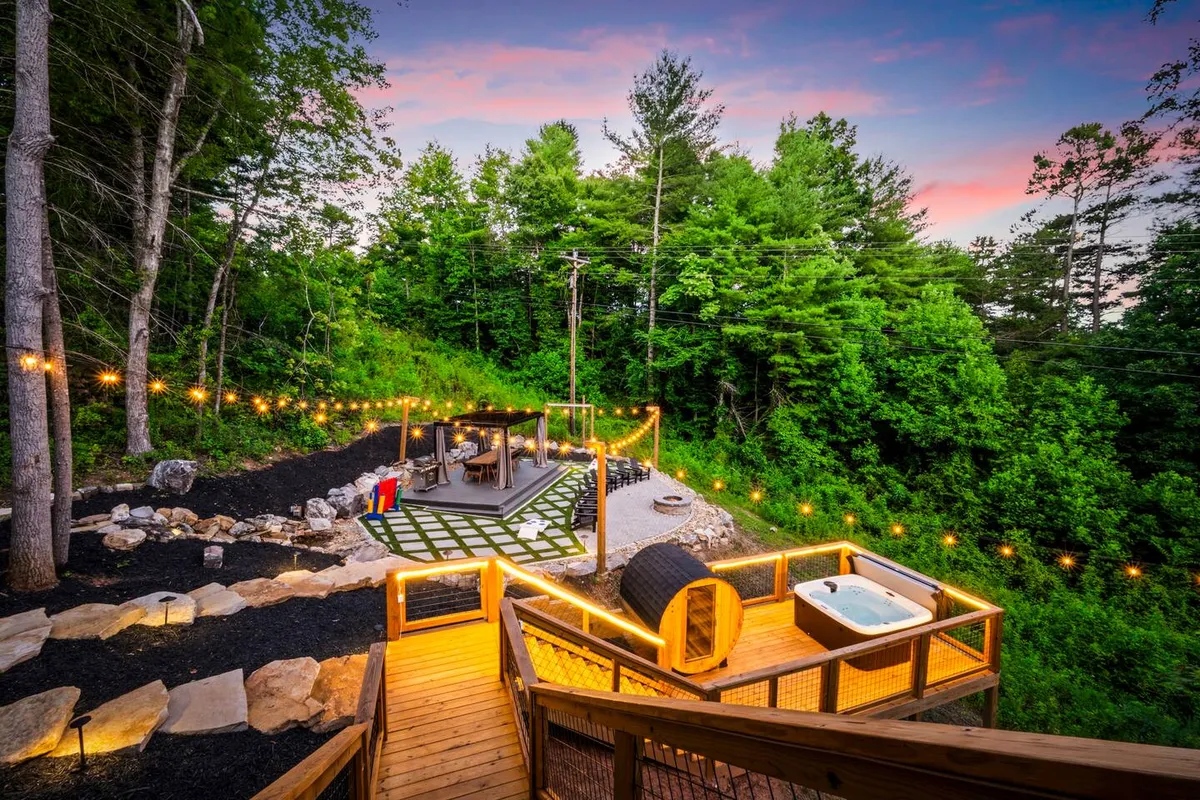 A hot tub sits on a wooden deck adorned with string lights, creating a welcoming space for relaxation. The area includes a pathway leading through the natural surroundings, highlighting a seamless connection to the outdoors. The ambiance is tranquil, ideal for unwinding or enjoying stargazing at night.