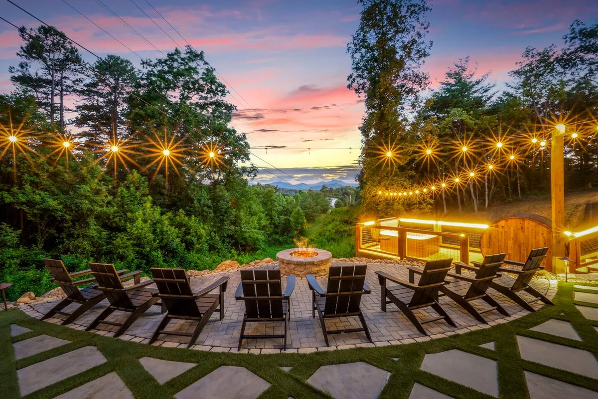 The image shows a cozy outdoor fire pit surrounded by Adirondack chairs, draped with glowing string lights, set against a breathtaking mountain backdrop. Guests are pictured gathering around the fire, sharing laughter, making s'mores, and soaking in the starlit sky, highlighting the property's inviting communal space and spectacular natural views.