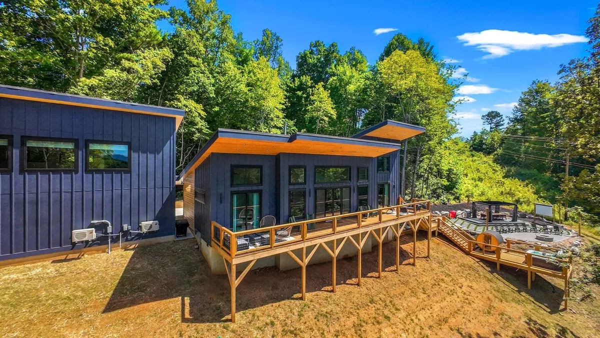 This exterior view of Starlit Lux Retreat AVL showcases its modern architecture and large wooden deck, surrounded by lush trees. The deck is set up for outdoor dining, relaxation by the fire pit, and gatherings under the starlit sky, epitomizing the luxury and comfort described in the narrative.