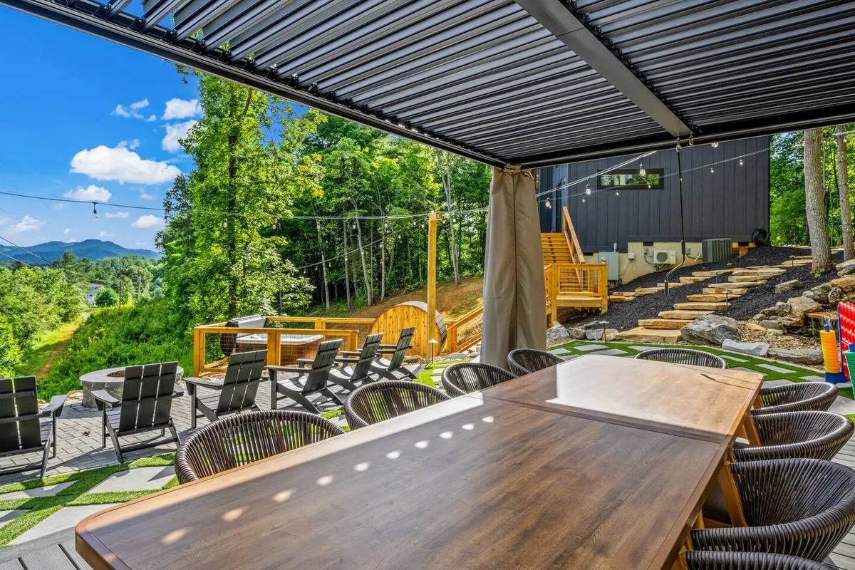 This image captures a modern outdoor dining area with a large wooden table, surrounded by lush greenery and set against impressive mountain views. The sleek architecture and comfortable seating invite guests to gather for meals, celebrations, or evening conversations.