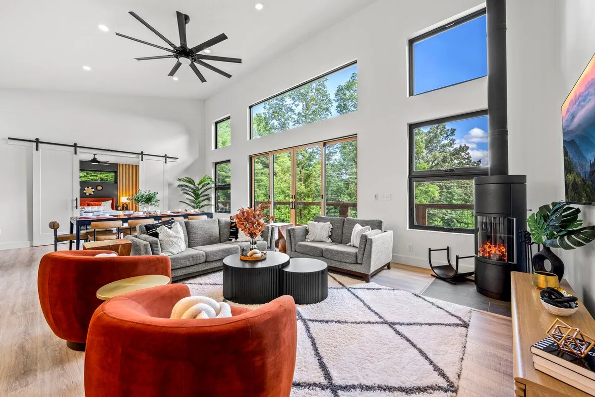 A spacious living room with a contemporary design, featuring large windows that offer beautiful outdoor views. The room includes a modern fireplace, comfortable seating arranged for conversation, and decorative plants, all bathed in natural sunlight.